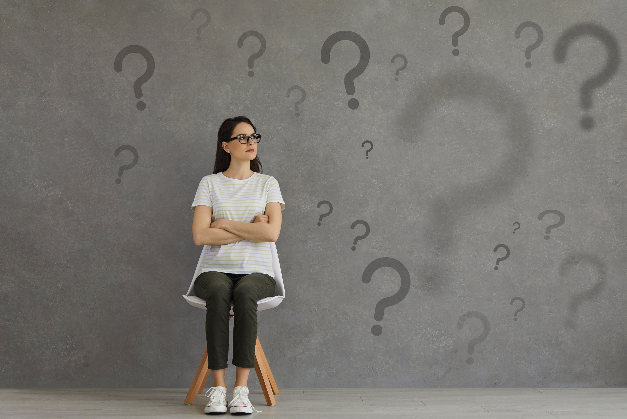 Confused Woman Sitting on Chair and Looking at Lots of Question Marks on Grey Background